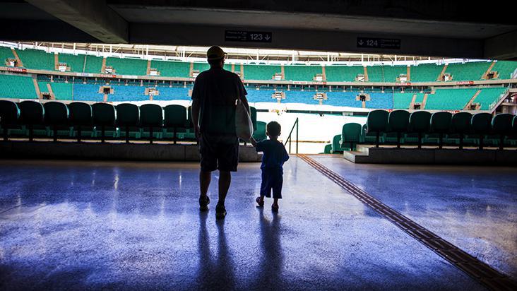 Imagem de um estádio de futebol, com cadeiras verdes e um homem e um menino de costas entrando nesse estádio