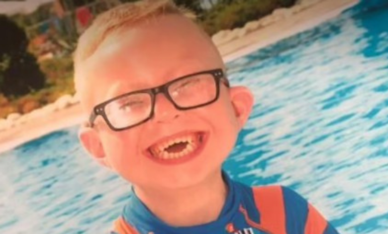 Na foto está o menino Logan Jones, em frente a uma piscina, usando óculos e com um lindo sorriso no rosto.
